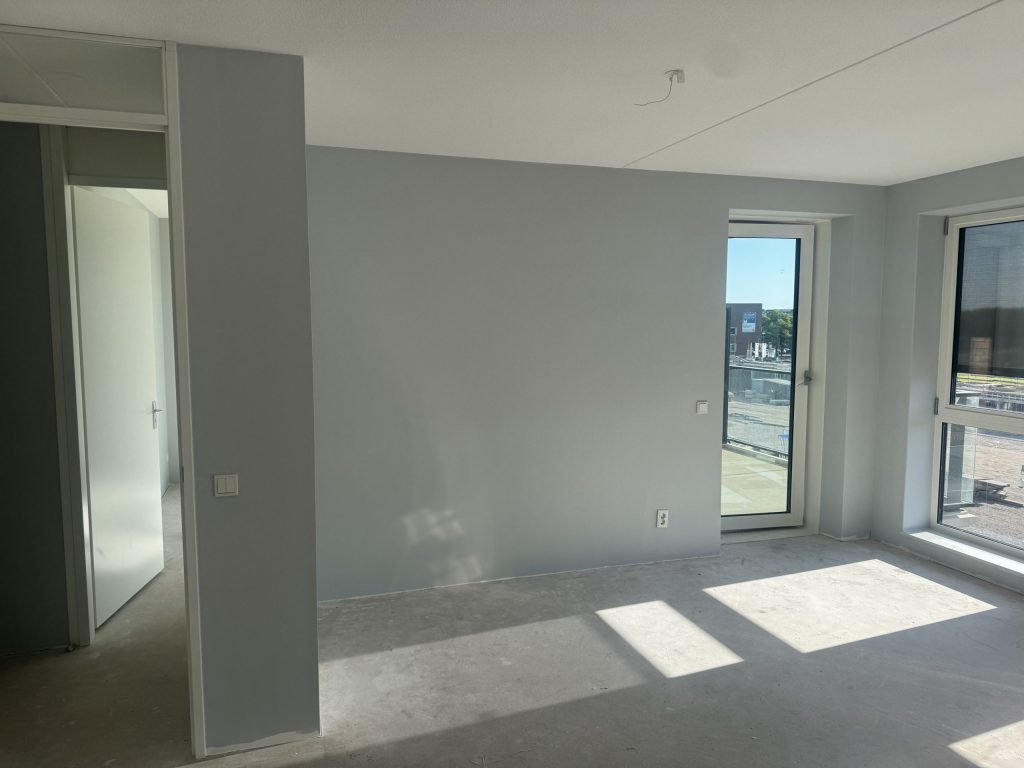 Lichtgrijs gesausde woonkamer in een nieuw appartement met een balkondeur en uitzicht op een bouwterrein