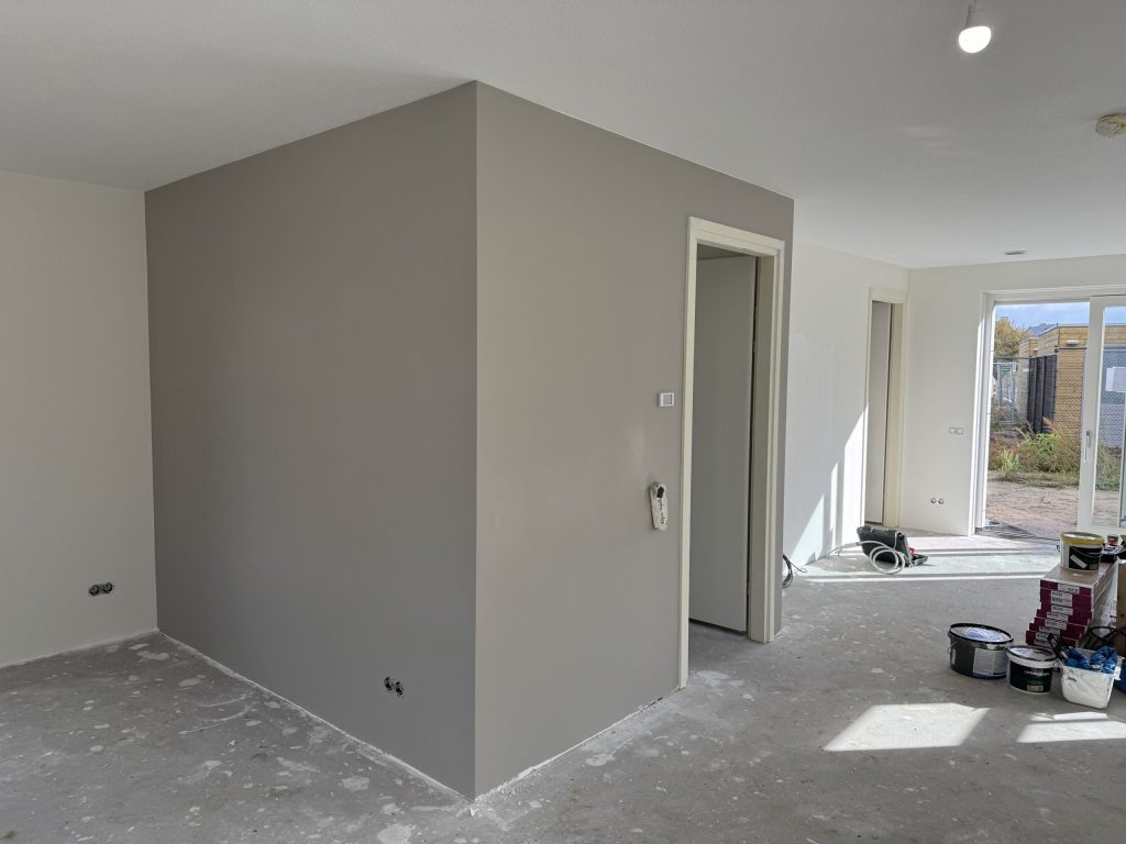 Nieuwbouwwoning met een taupe gesausde wand rond de meterkast, verfemmers op de betonvloer en tuindeuren achterin