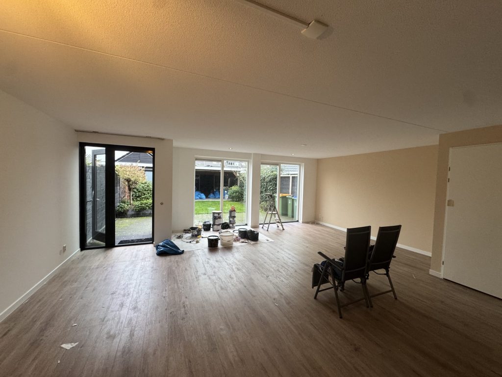 Ruime woonkamer met lichte wanden, een houten vloer, tuindeuren en materiaal van de behanger bij het raam