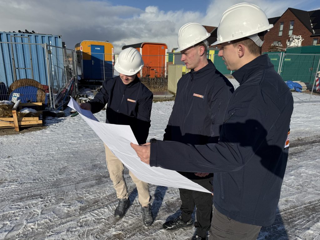 Drie medewerkers van Bouwsector Nederland met witte bouwhelmen bekijken een bouwtekening op een besneeuwd nieuwbouwterrein